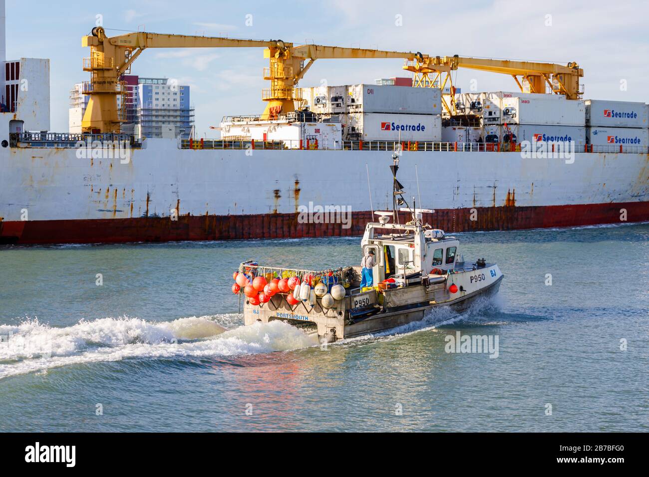 A small traditional fishing is dwarfed as it passes by a large container ship in Portsmouth Harbour, Portsmouth, Hampshire, south coast England Stock Photo