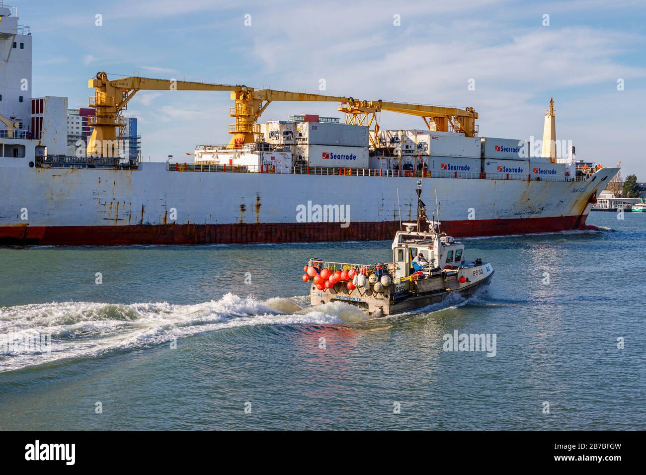 A small traditional fishing is dwarfed as it passes by a large container ship in Portsmouth Harbour, Portsmouth, Hampshire, south coast England Stock Photo