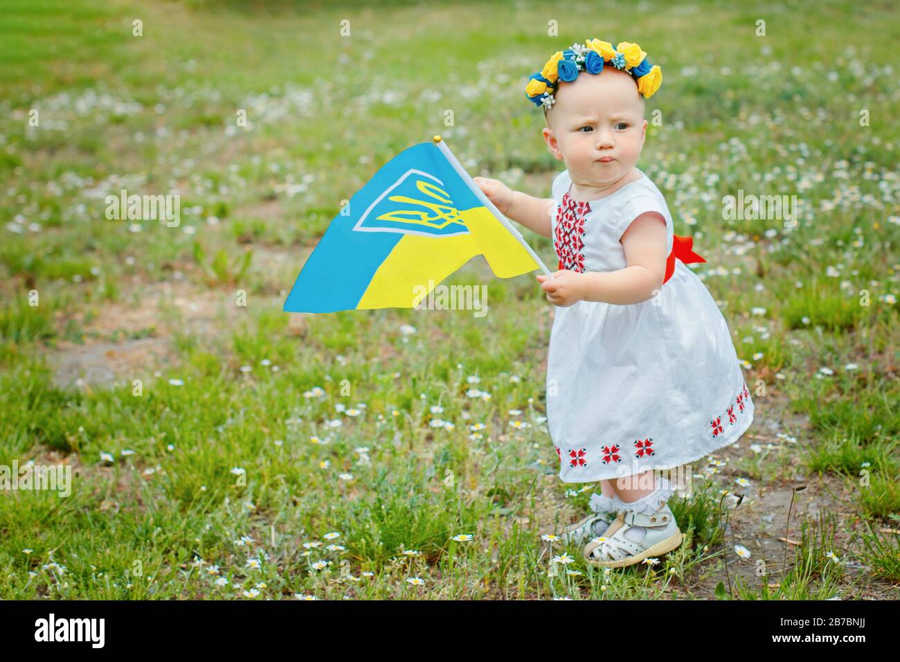 little girl with Ukrainian embroidery and wreath with yellow and blue flowers is carried by Ukrainian flag. Happy baby kid on Independence Day of Ukra Stock Photo
