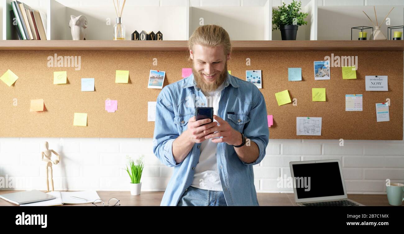 Smiling millennial man standing at home office workspace using cell phone. Stock Photo