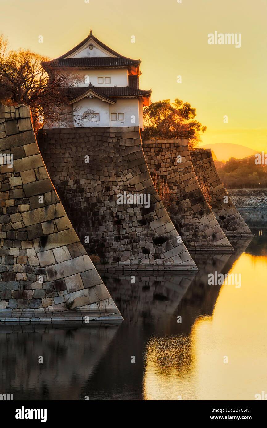 Bastions of historic castle stone walls growing from water moat in Osaka city of Japan lit by rays of rising sun. Stock Photo
