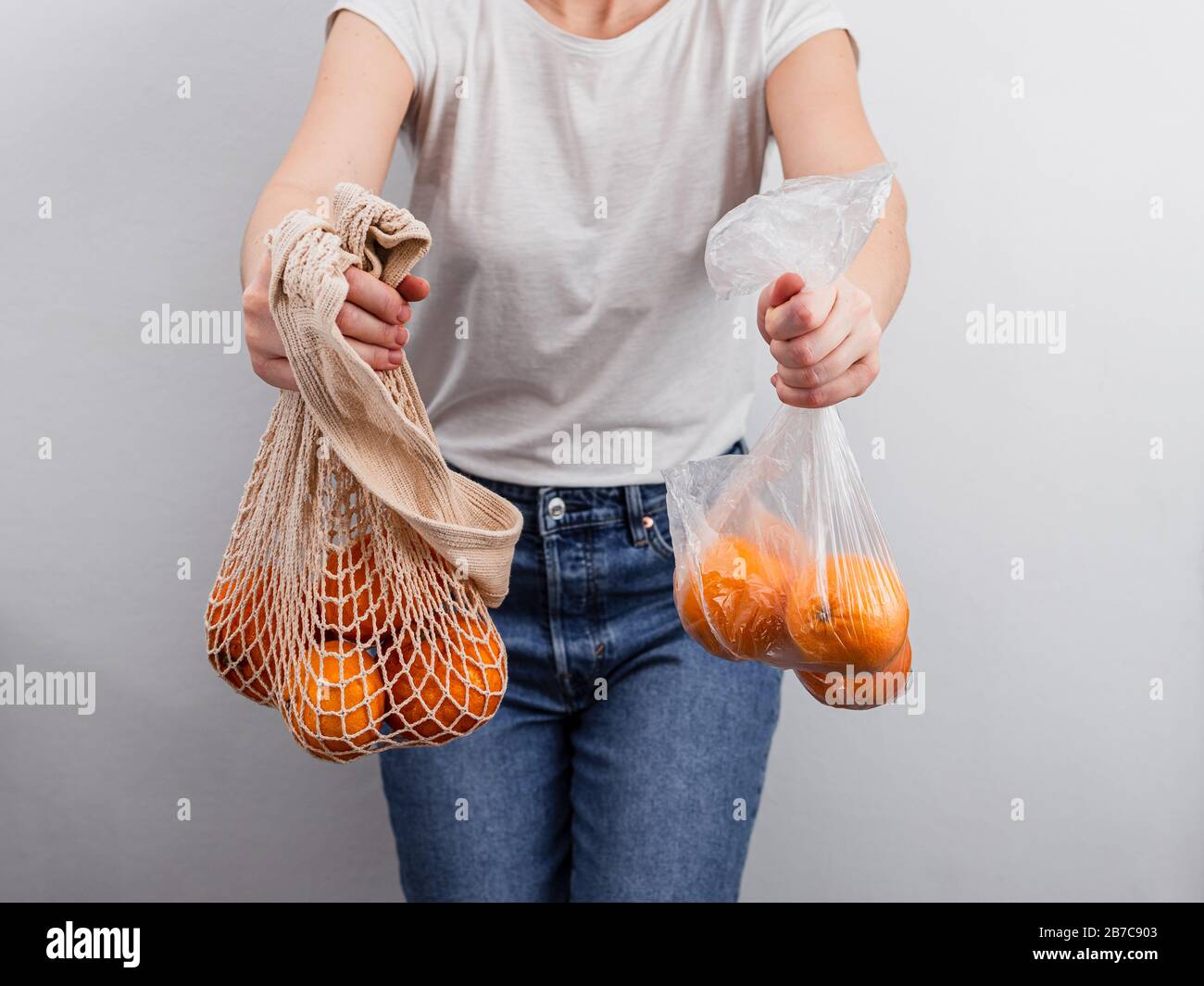 Oranges fruits in plastic bag and oranges in textile mesh bag in female hands near white wall