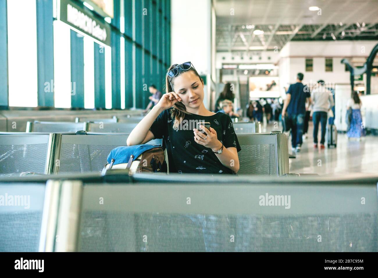 A tourist or student is waiting for a boarding announcement at a Milan airport in Italy to leave the country. She uses the phone, reads messages, or w Stock Photo