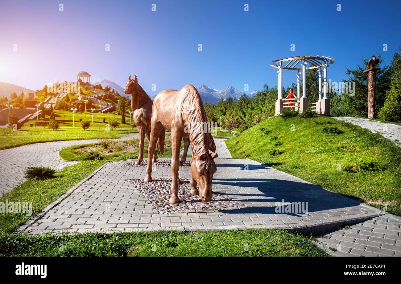 Woman in orange dress in the park with horse statue and mountains in Almaty, Kazakhstan Stock Photo