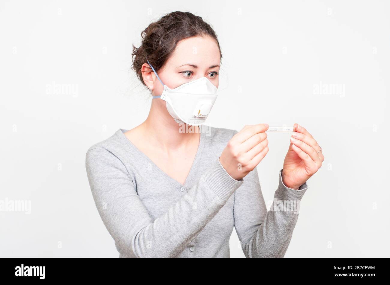 caucasian brunette woman in protective medical mask on face SARS-CoV-2, Coronavirus, COVID-19. with mercury thermomether. white background. Stock Photo