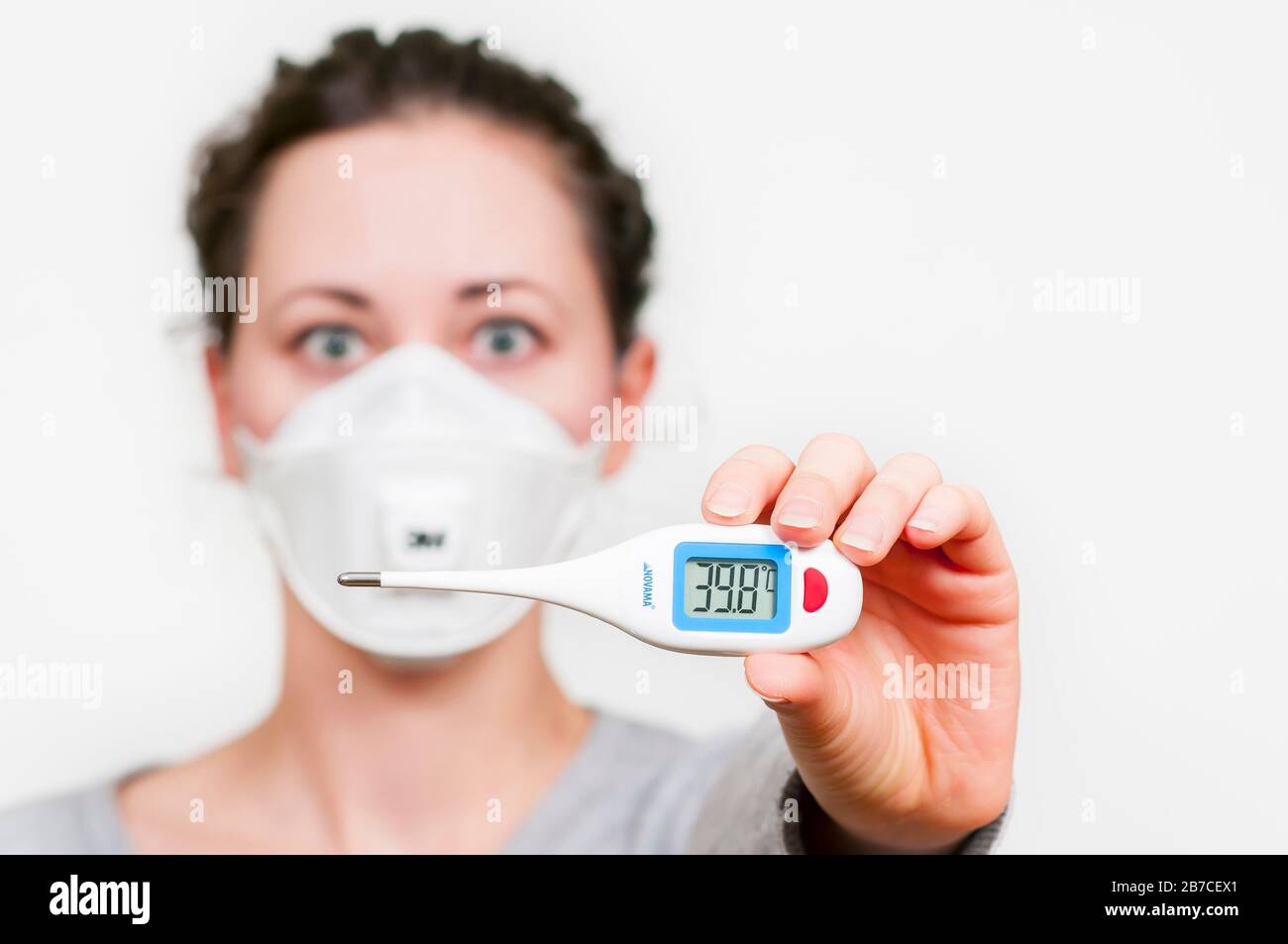 caucasian brunette woman in protective medical mask on face SARS-CoV-2, Coronavirus, COVID-19. with electric, digital thermomether. white background. Stock Photo