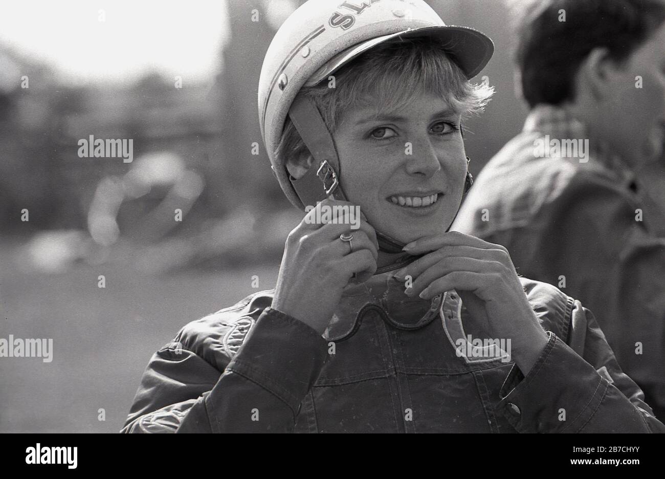 1980s, historical, close-up of a female rider adjusting her hard protective hat before taking part in a harness race, a sport involving steering or riding a horse from a cart, sometimes referred to as a pony and trap, England, UK. Stock Photo