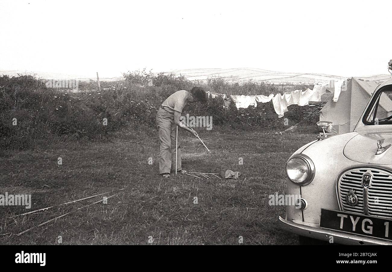 Camping at the coast 1950s style, male camper in field next to his Austin motorcar sorting out the poles of his tent, next to s neighbour with washing hanging on a washing line, England, UK. H Stock Photo