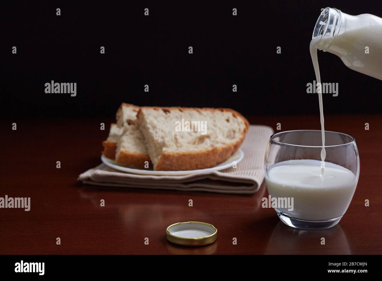 Kefir from a bottle flows into a glass. Bread in the background. Dark background and brown wooden table. Stock Photo