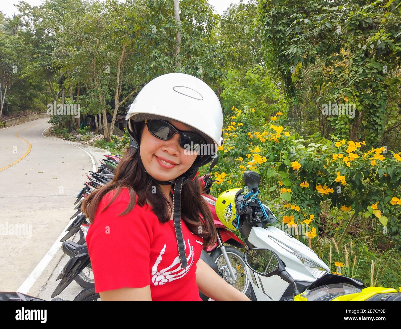 Young Thai lady wearing an helmet. Behind her, a green motorcycle and a tropical background Stock Photo