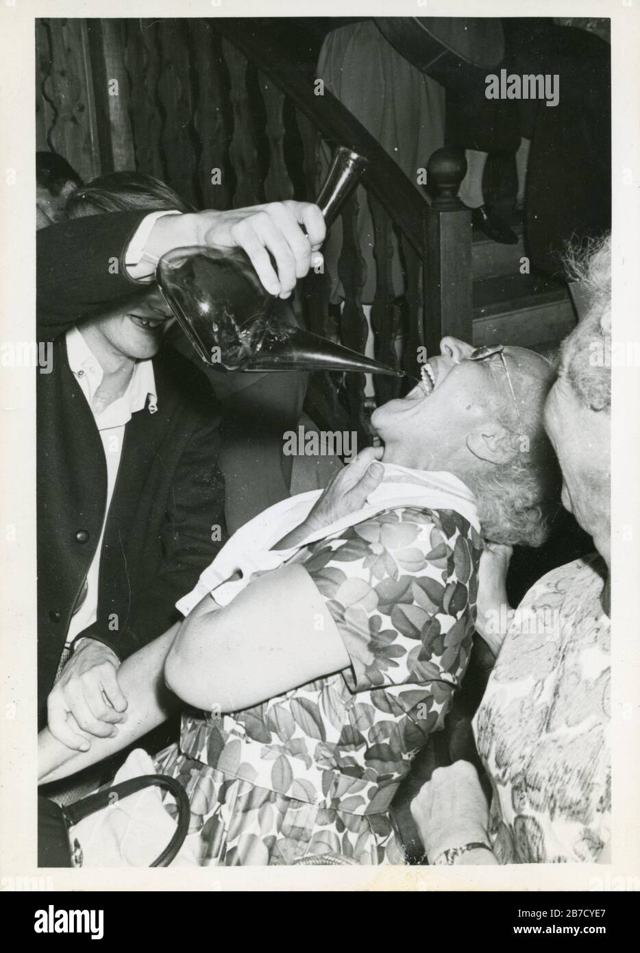 1960s black and white snapshot of an elderly lady wearing a flowery frock being forcefed wine or sangria from a porron by a young man whilst another elderly lady looks on in the foreground. Vernacular photography found photo. Photographer unknown (Richard Bradley collection) Stock Photo