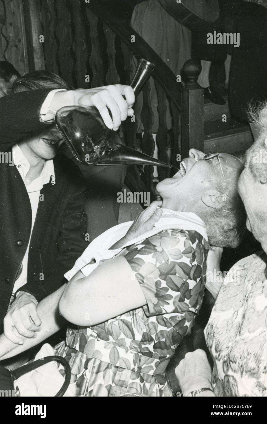 1960s black and white snapshot of an elderly lady wearing a flowery frock being forcefed wine or sangria from a porron by a young man whilst another elderly lady looks on in the foreground. Vernacular photography found photo (Same as Alamy image #2B7CYE7 without white border). Photographer unknown (Richard Bradley collection) Stock Photo