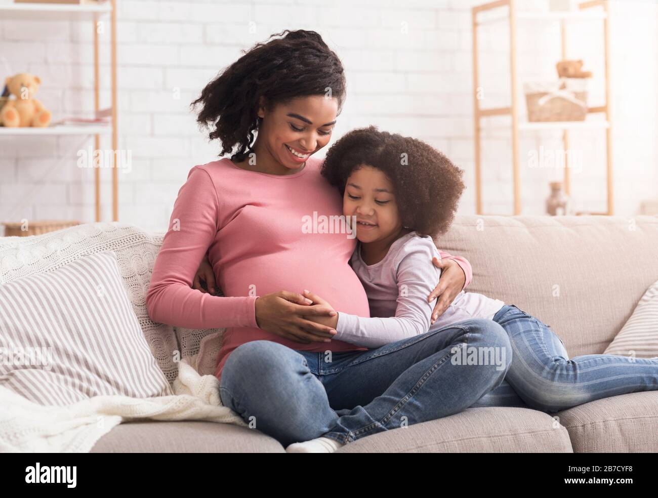 Affectionate Little Daughter Tenderly Touching Belly Of Pregnant Mom On Sofa Stock Photo