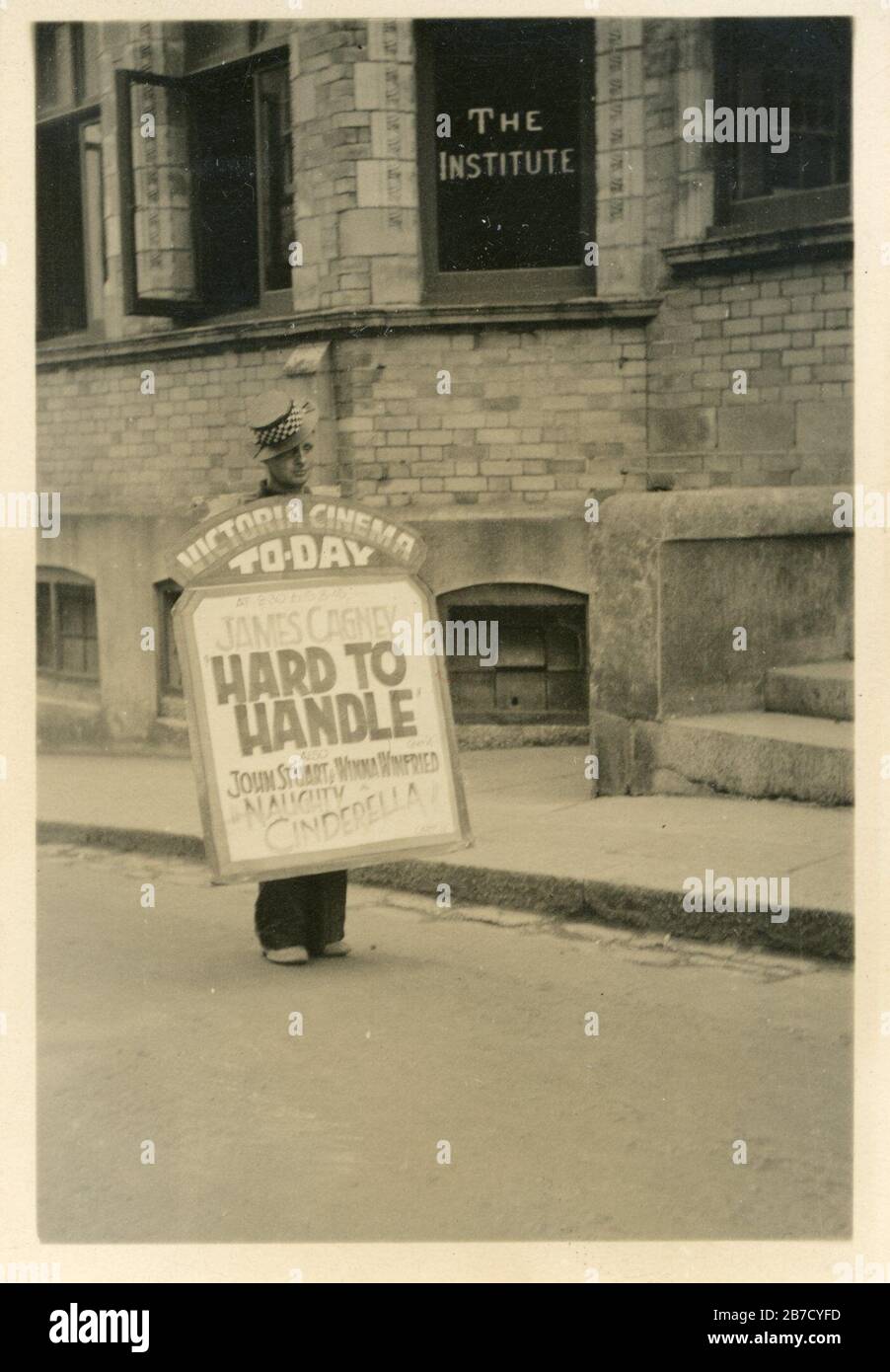 Vintage snapshot photo of a man with a sandwich board walking down the  street to advertise the 1933 James Cagney film 'Hard To Handle' which is  showing at the Victoria Cinema (also
