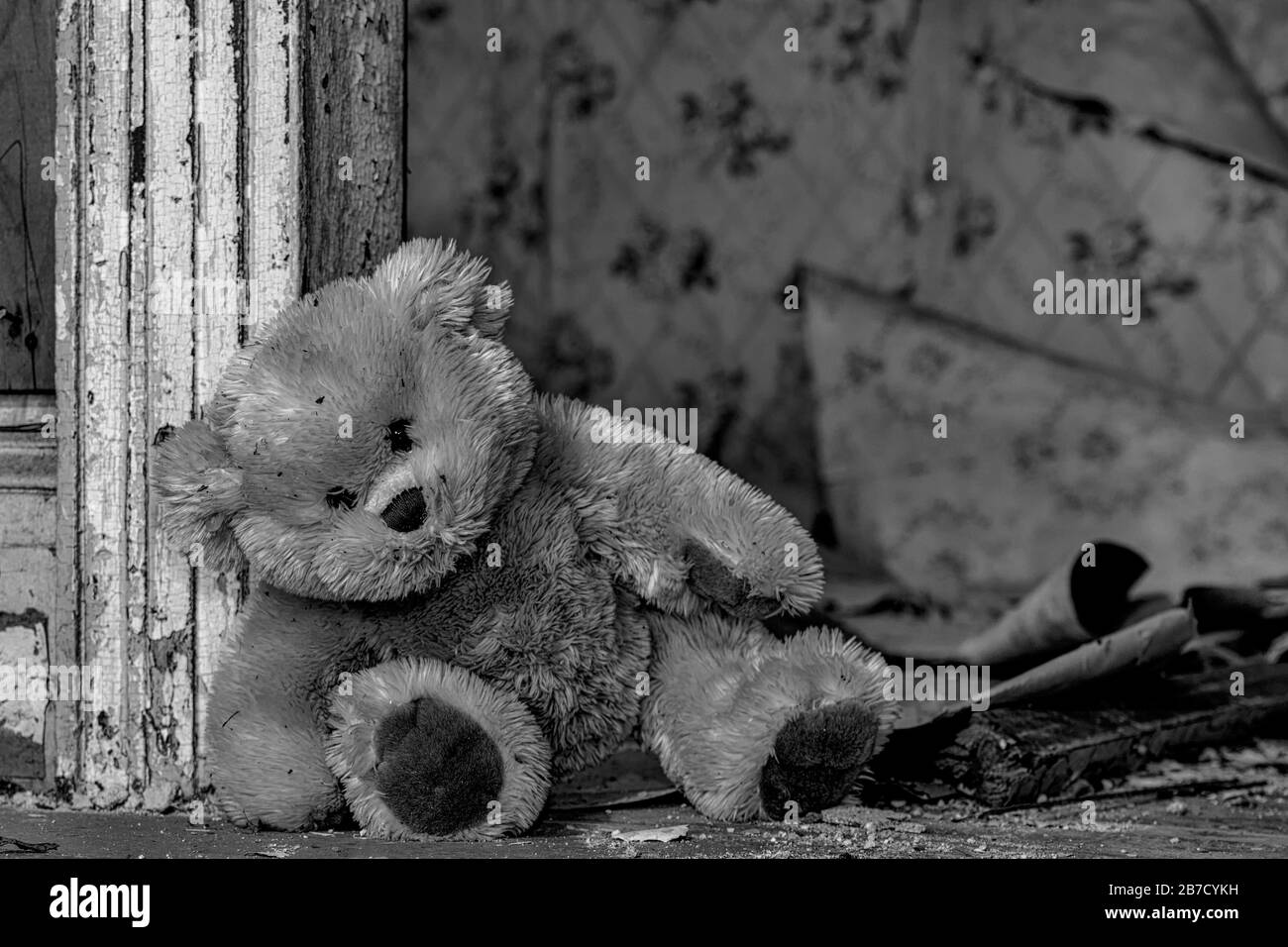 A dirty teddy bear leaning against a door frame in an old, abandoned house. Debris and assorted wallpaper peeling from the wall in the background. B&W Stock Photo