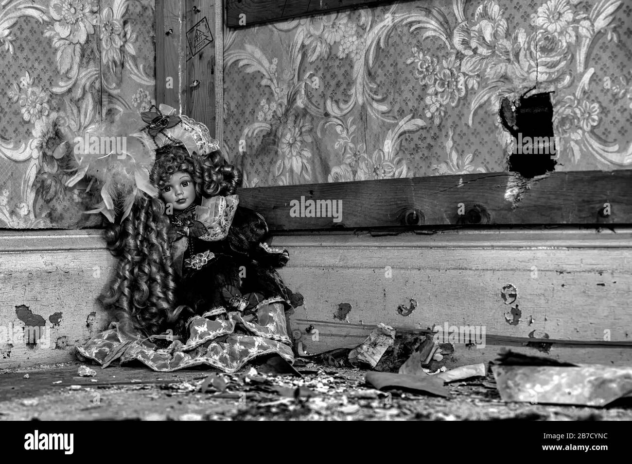 An old dall sitting ina corner in an abandoned house. There is a hole in a wall, and the floor is littered with dirt and debris. Black and white. Stock Photo