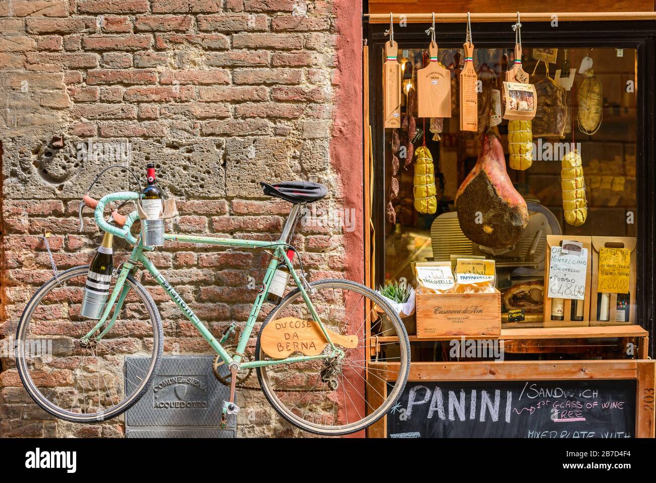A bike hanging on the brick wall by the entrance of Alimentari Du' Cose Da Berna, a  delicatessen selling local specialities in Siena, Tuscany, Italy. Stock Photo