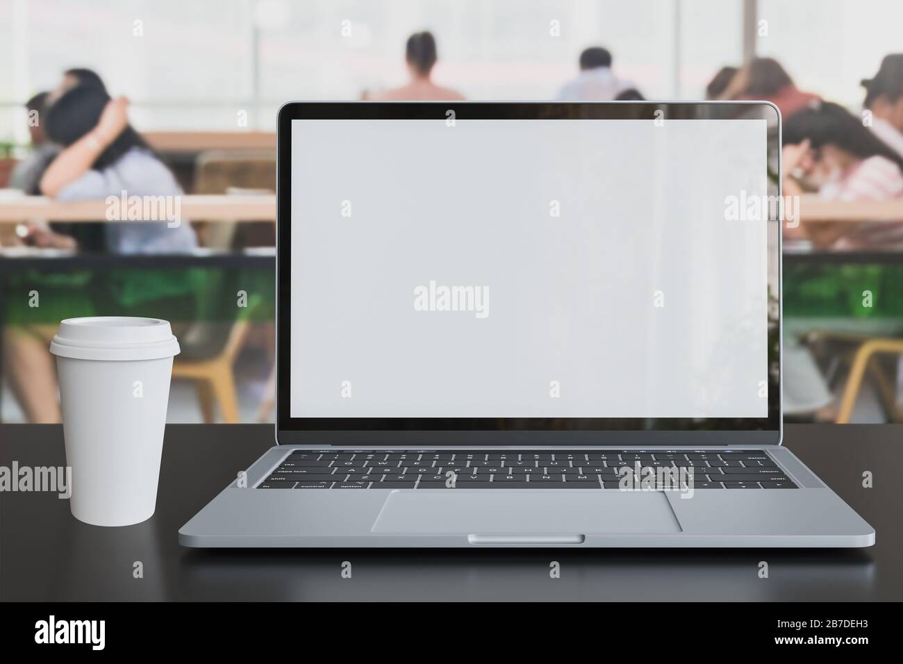 Laptop blank white screen placed with white coffee cup on table in co-working space with Defocused customer at cafe coffee shop restaurant blurred for Stock Photo
