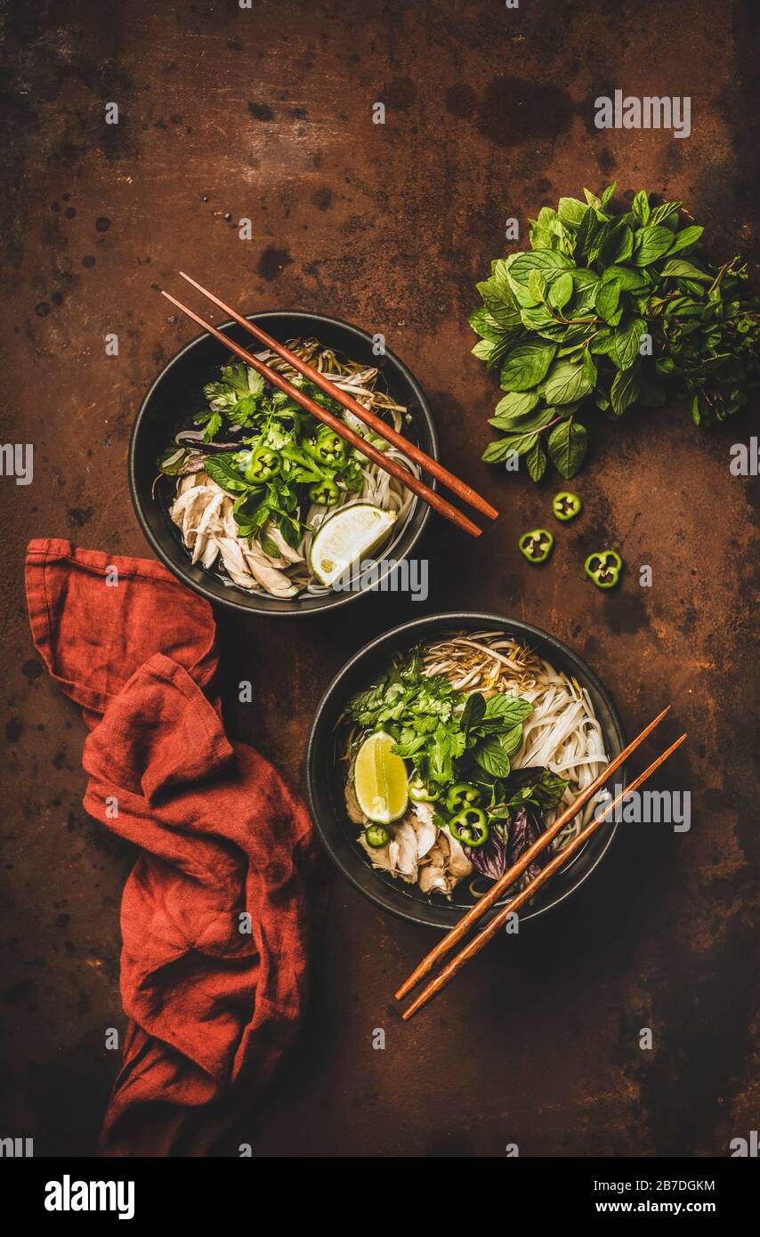 Asian cuisine lunch. Flat-lay of Vietnamese rice noodle chicken soup Pho Ga with cilantro, soy sprouts, fresh greens, lime in black bowls with chopsti Stock Photo