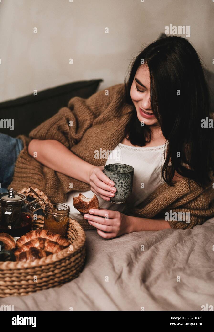 Young smiling brunette woman in comfortable home clothing having breakfast in bed with tea and croissants at home. Comfortable lifestyle concept Stock Photo
