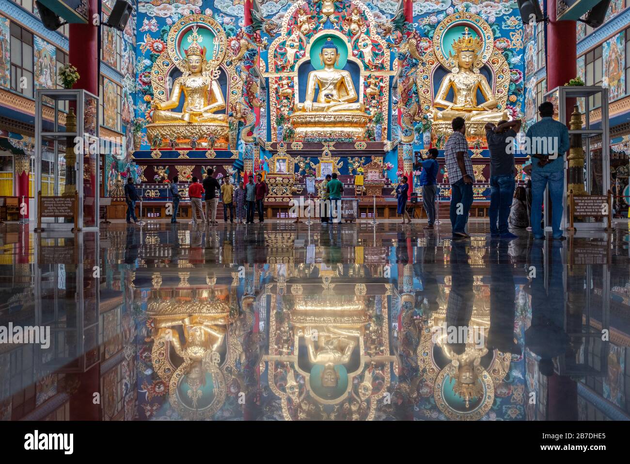 Bylakuppe, Karnataka, India - March 15, 2018: Visitors in the main temple of at the Namdroling Tibetan, Monastery with three buddhas reflecting on a s Stock Photo