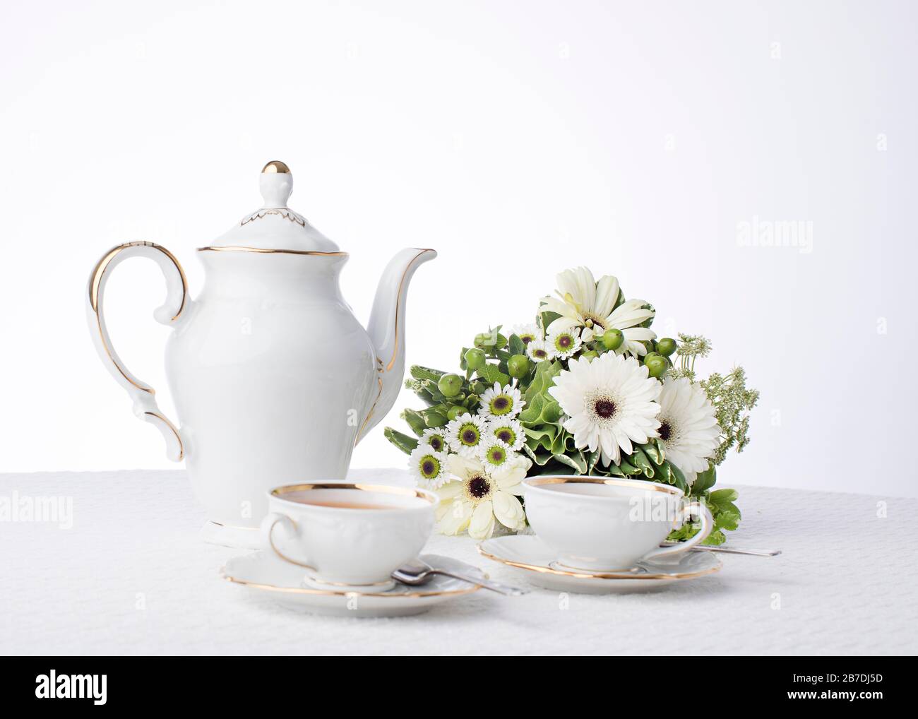 Fresh white assorted daisies and green leaves decorate the table for tea time with antique cups and saucers on white background with room Stock Photo