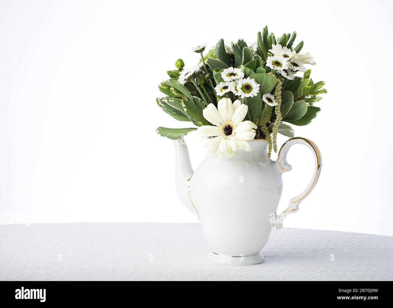 White tablecloth and white china tea pot as a vase sets off the greenery and white flower petals in the floral centerpiece Stock Photo