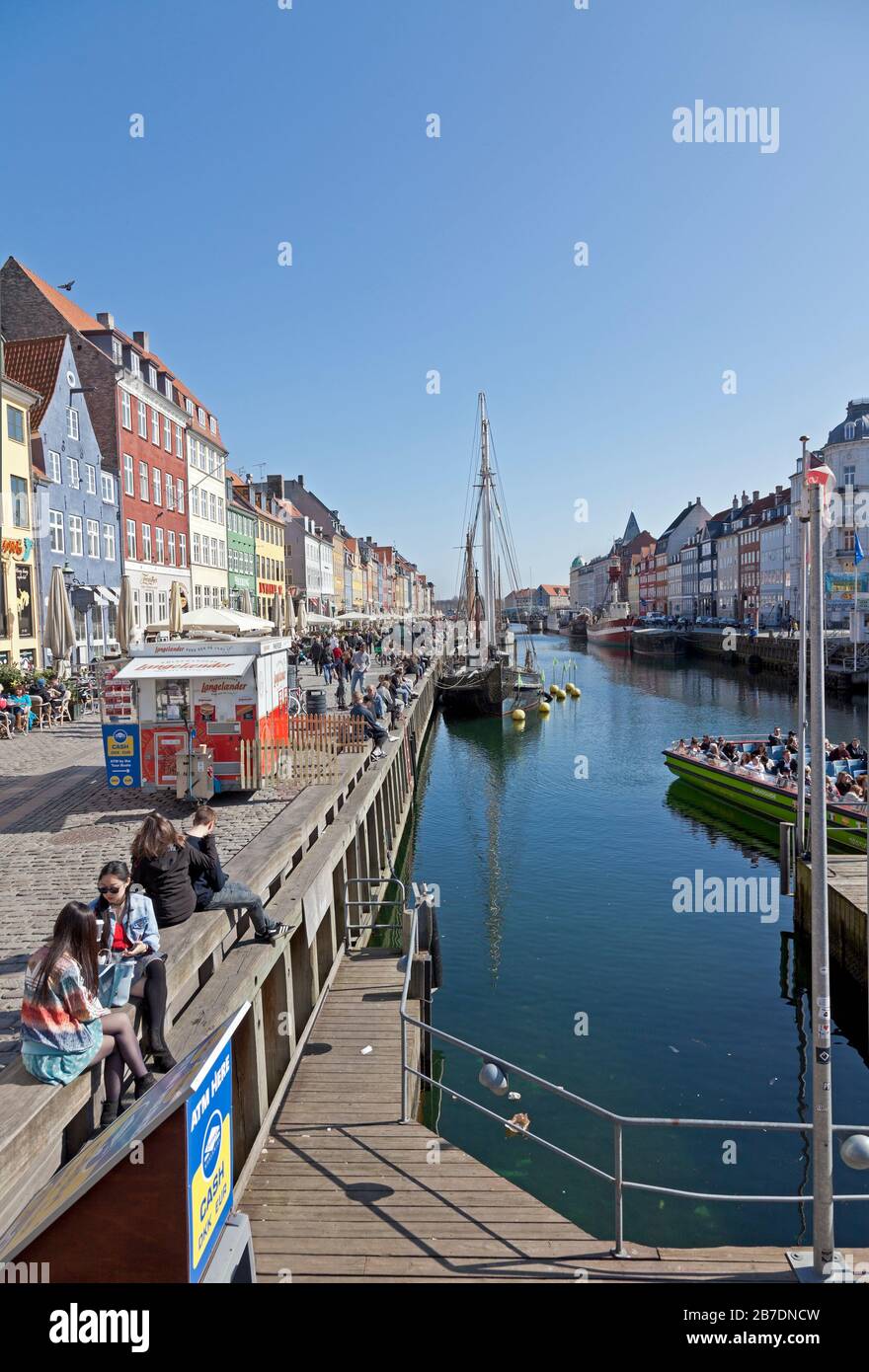 View along the Nyhavn canal on a sunny and nearly warm spring day. Copenhagen, Denmark. Copenhageners and tourists enjoy the sunshine. Stock Photo