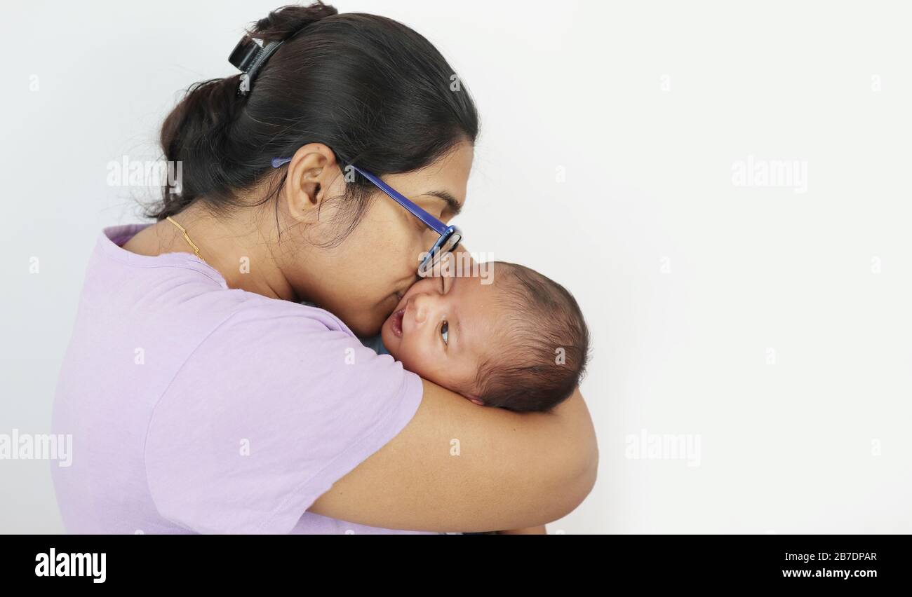 a young mother in spectacles kissing her infant baby boy in her lap in solid grey background with space for text. mothers day concept photo. Stock Photo