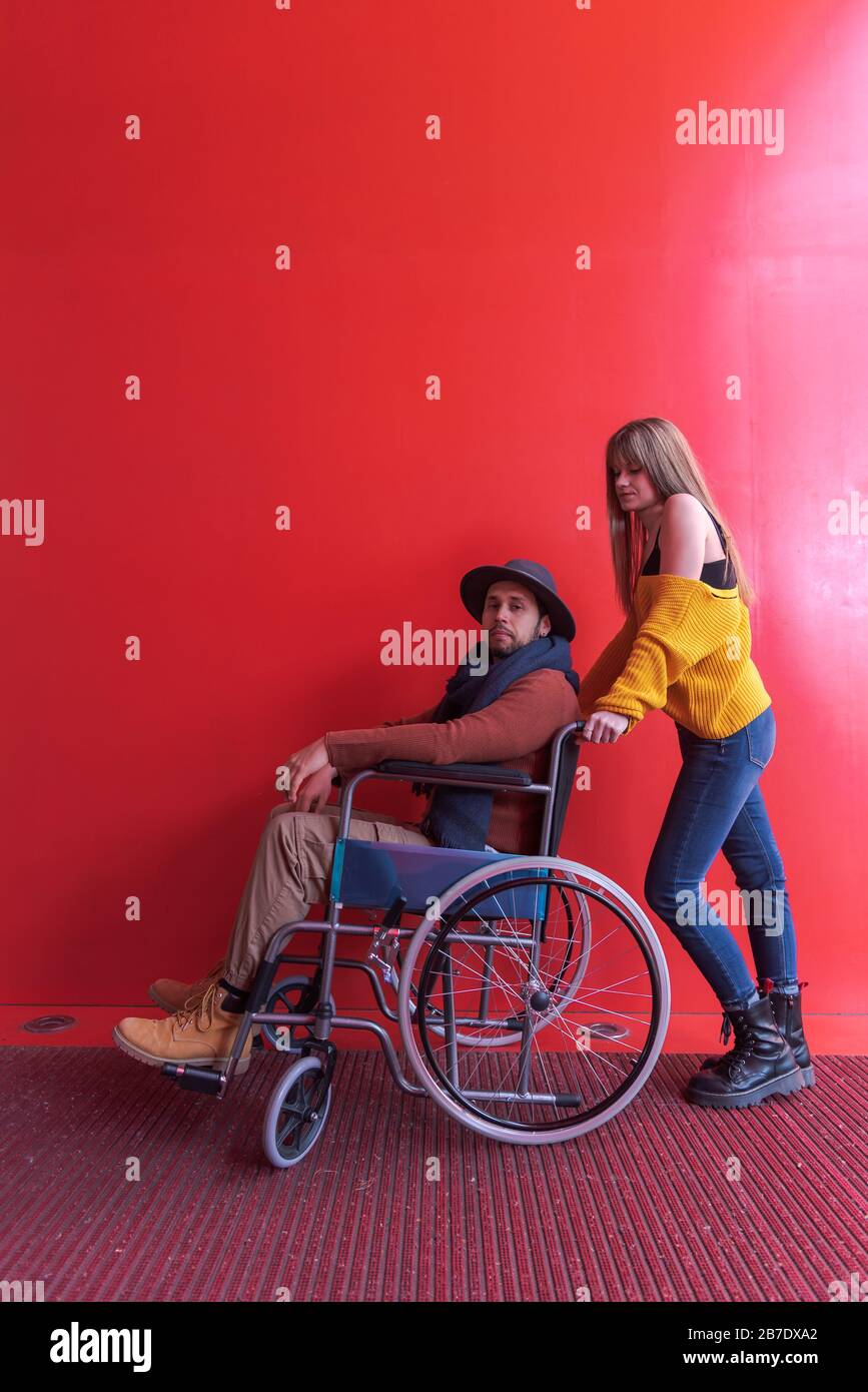 Disabled in a wheelchair pushed by a young blonde girl in front of a red background wall Stock Photo