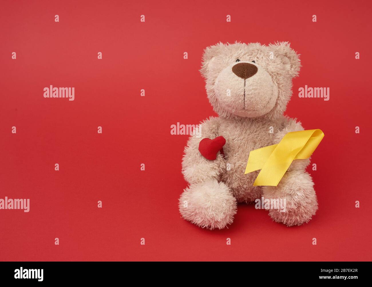 Cute brown teddy bear holds a silk yellow ribbon in the shape of a loop on a red background, concept of the fight against childhood cancer, problem of Stock Photo