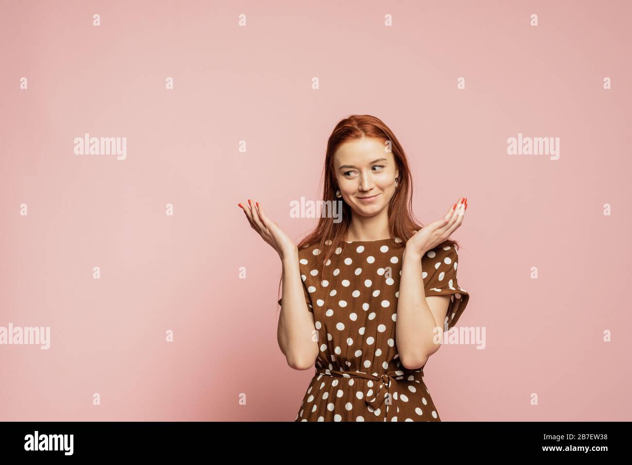 Confused and puzzled woman in the studio on a pink background. A girl in a beautiful brown dress shows the emotion of not understanding. I dont know. Stock Photo