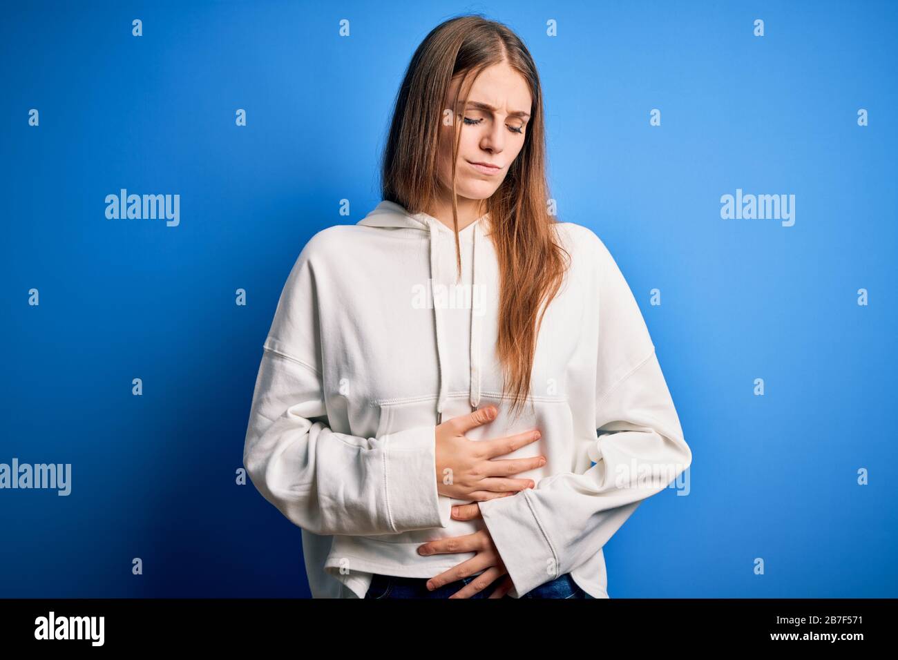 Young beautiful redhead sporty woman wearing sweatshirt over isolated blue background with hand on stomach because indigestion, painful illness feelin Stock Photo