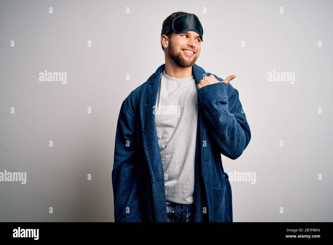 Young blond man with beard and blue eyes wearing sleep mask and pajama smiling with happy face looking and pointing to the side with thumb up. Stock Photo