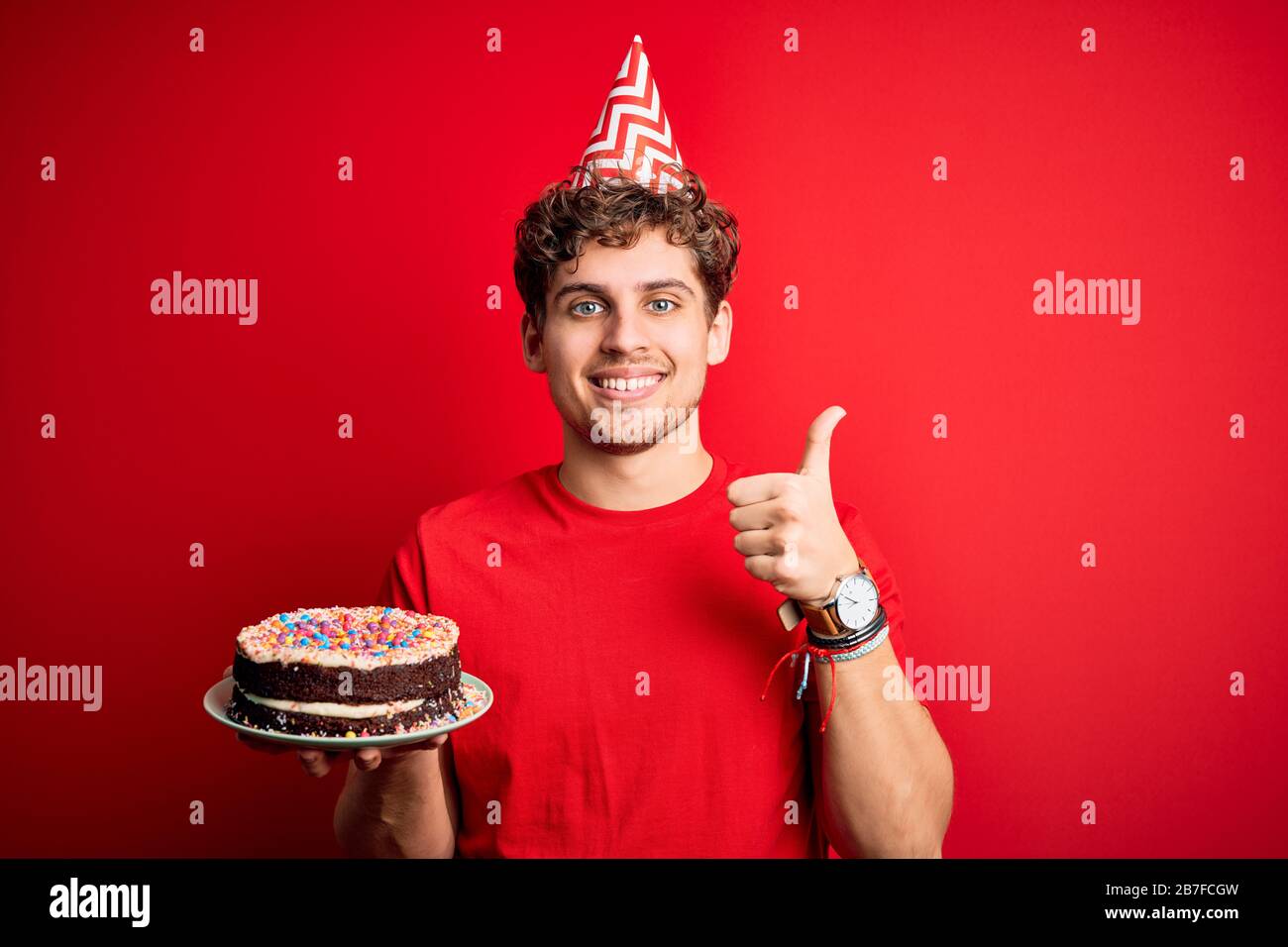 Young blond man with curly hair wearing birthday hat holding cake over ...