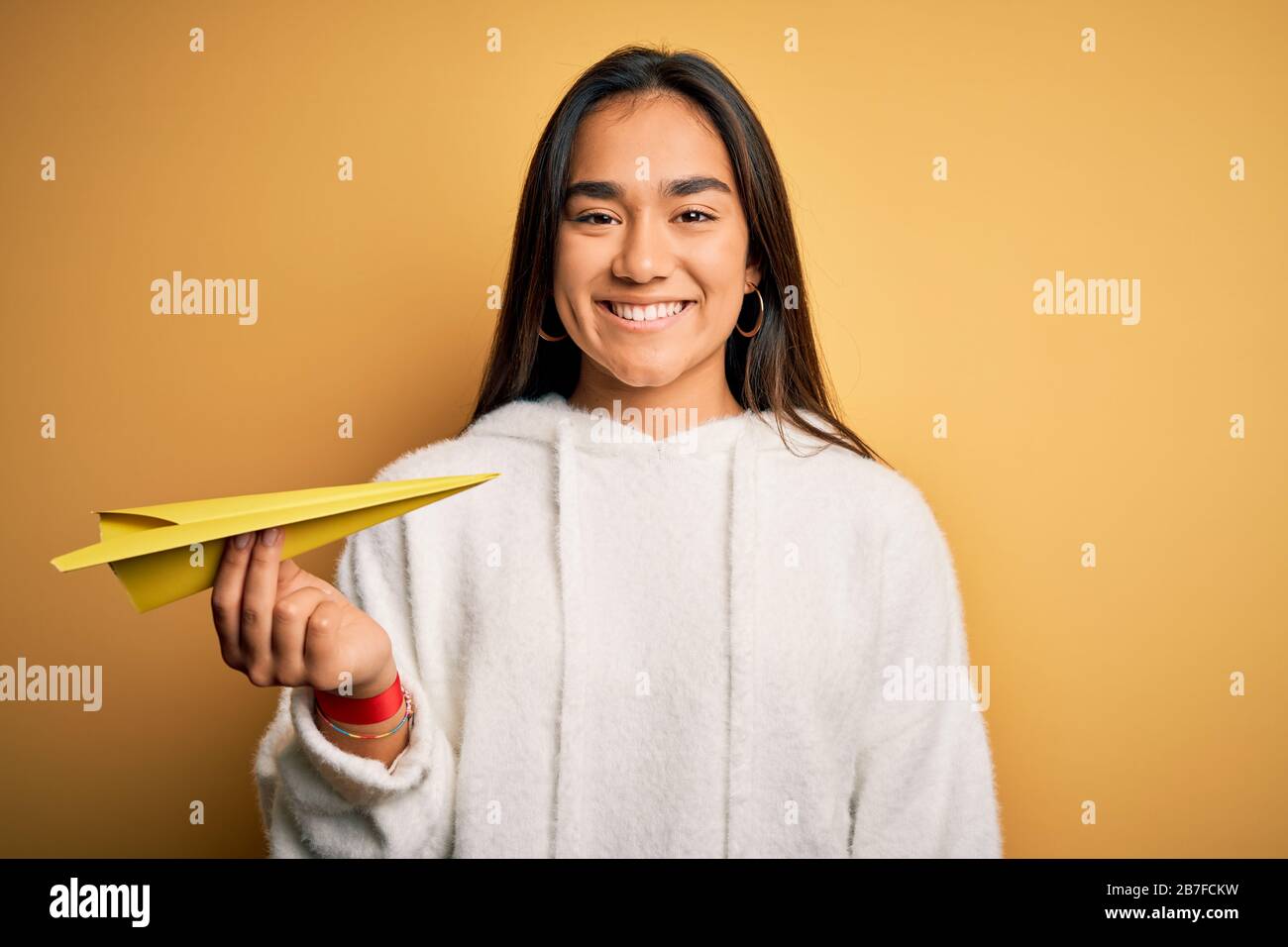 Young beautiful asian woman holding paper airplane standing over isolated yellow background with a happy face standing and smiling with a confident sm Stock Photo