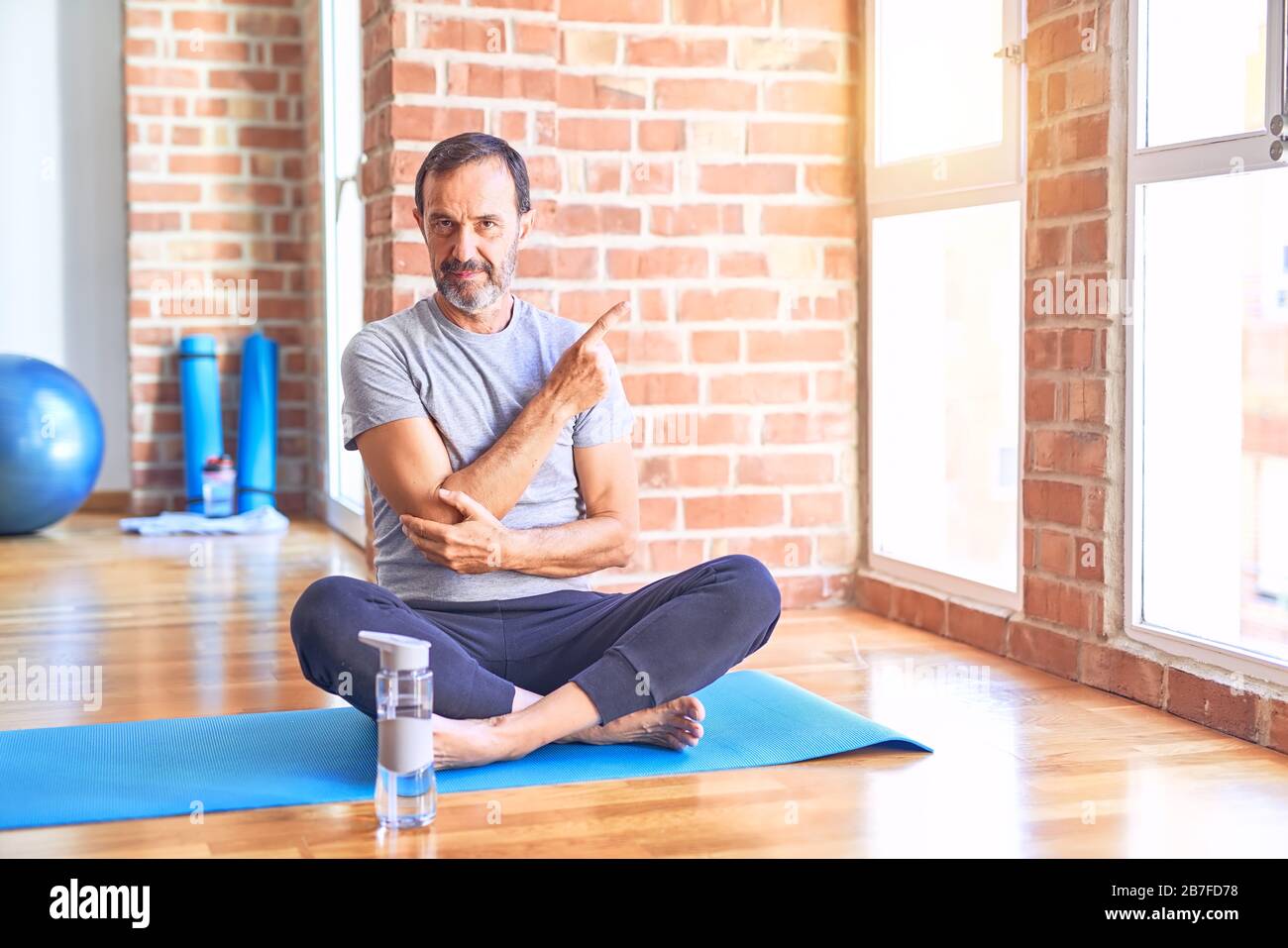 Middle age handsome sportman sitting on mat doing stretching yoga exercise at gym Pointing with hand finger to the side showing advertisement, serious Stock Photo