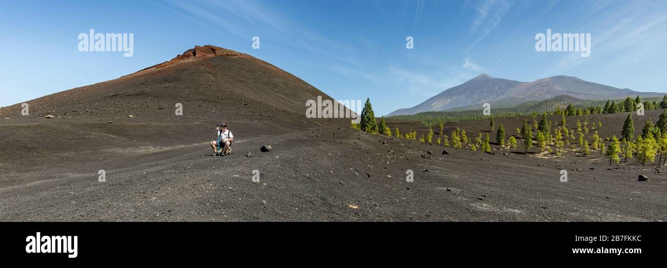 Super wide panoramic view of Volcano Arenas Negras and lava fields around. Bright blue sky and white clouds. Young traveler sits on a stone and rests. Stock Photo