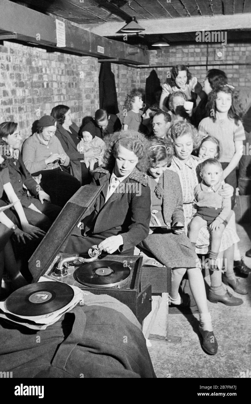 A young woman plays a gramophone in an air raid shelter in north London during 1940. A young woman places the gramophone needle on a record to bring some light relief to an air raid shelter, somewhere in north London. The rest of the shelterers appear to be enjoying her choice of music. In the background, one woman can be seen knitting, as others chat to pass the time. Stock Photo