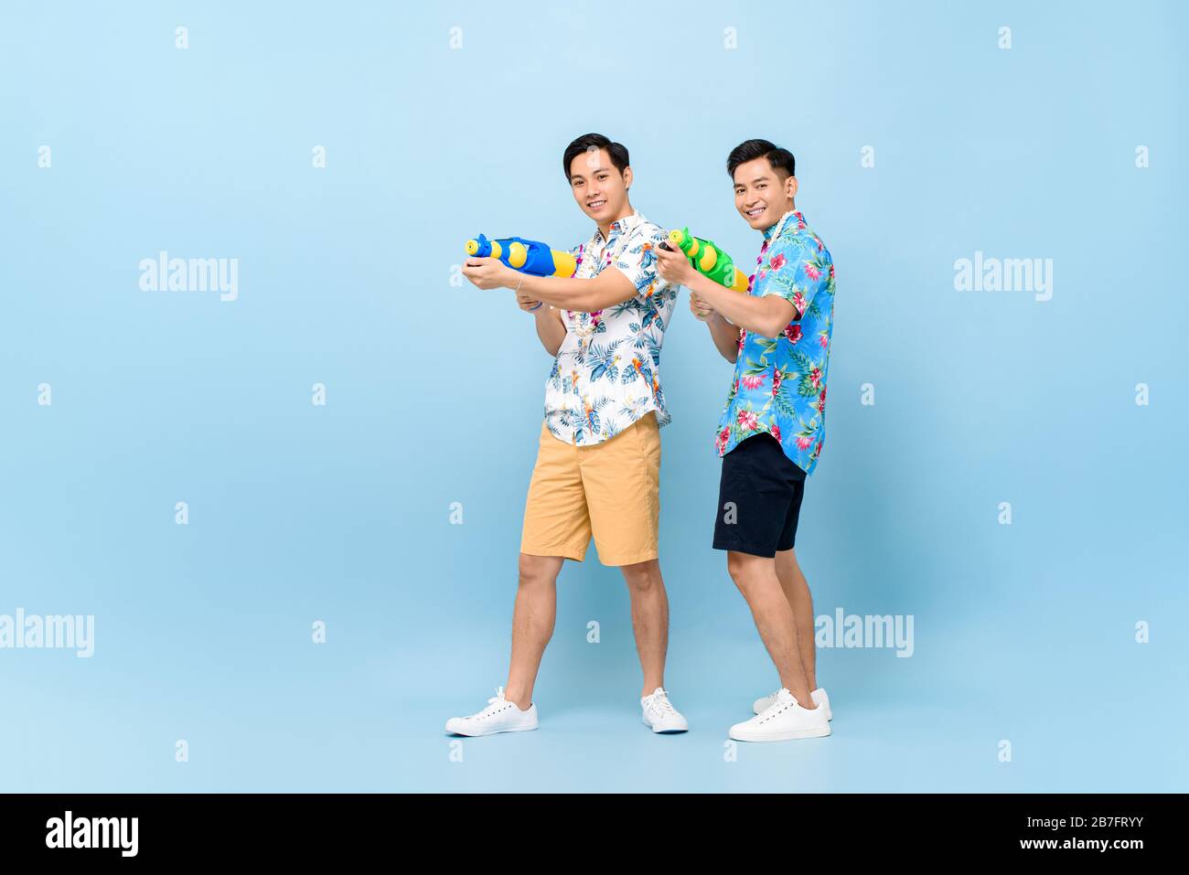 Smilng happy Asian male friends playing with water guns in blue isolated background for Songkran festival in Thailand and southeast Asia Stock Photo