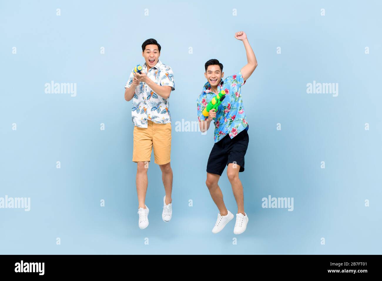 Smilng happy Asian male friends playing with water guns and jumping in blue isolated background for Songkran festival in Thailand and southeast Asia Stock Photo