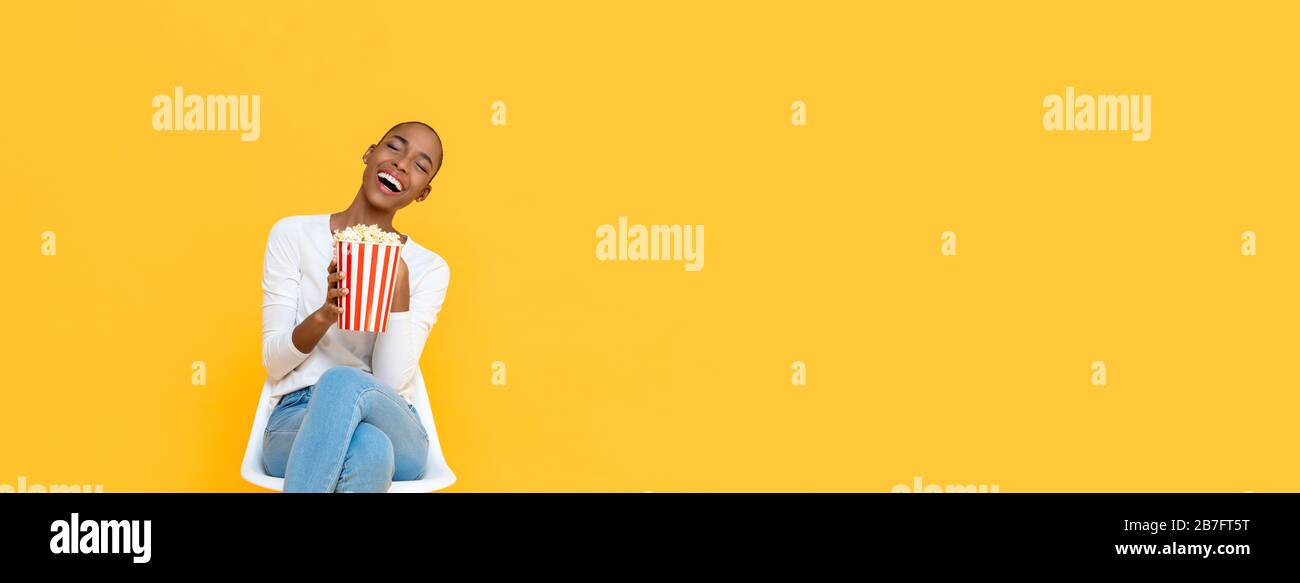 Happy young African American woman laughing while watching movie with popcorn in hand isolated on colorful yellow banner background with copy space Stock Photo