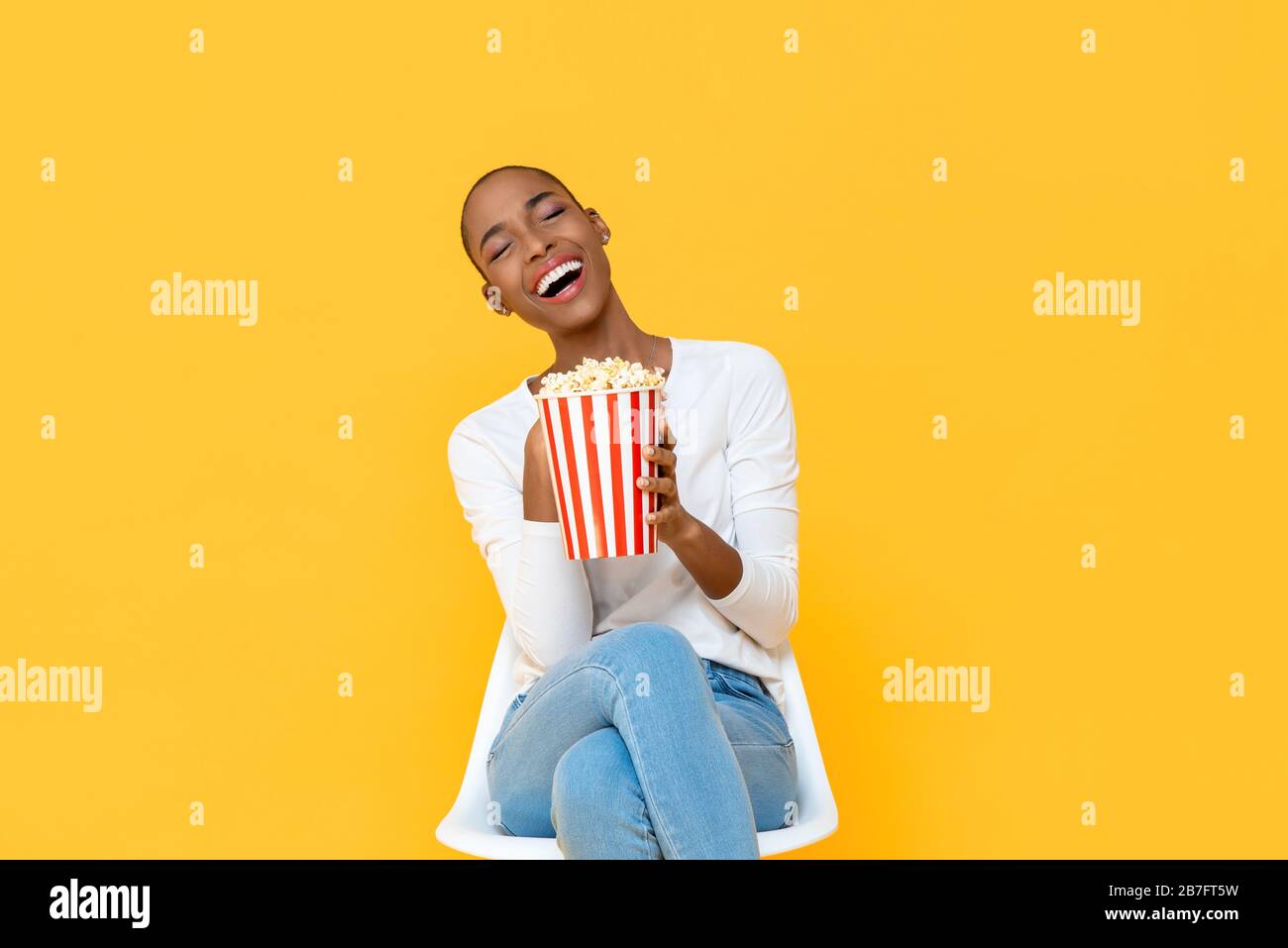 Happy young African American woman laughing while watching movie with popcorn in hand isolated on colorful yellow background Stock Photo