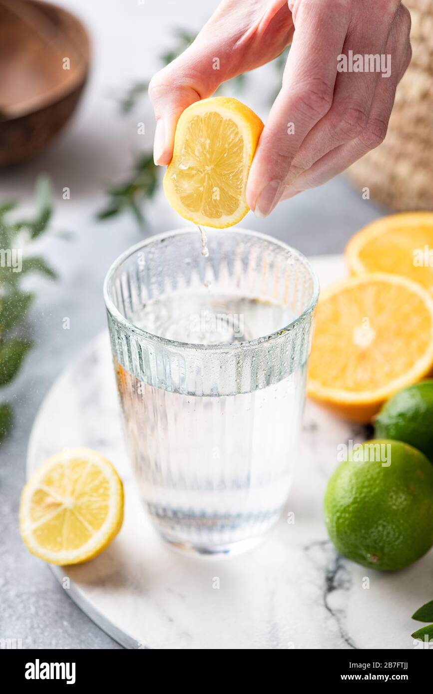 Lemon citrus water in glass. Woman hand squeeze lemon in glass of fresh clean water. Refreshing cold summer drink Lemonade Stock Photo