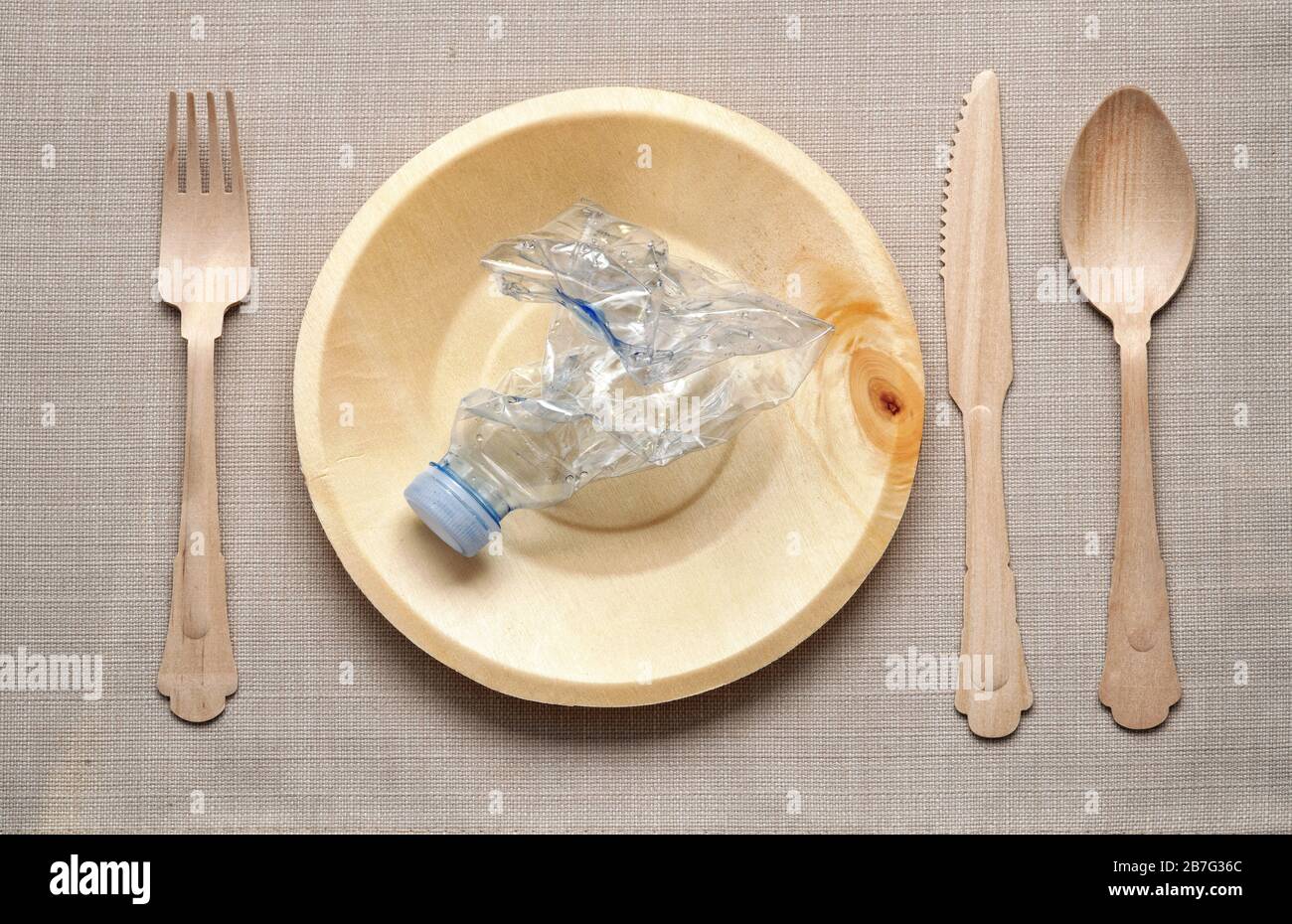 Eating plastic concept with table setting and a crumpled plastic bottle on a natural wood plate flanked by matching knife, fork and spoon viewed from Stock Photo