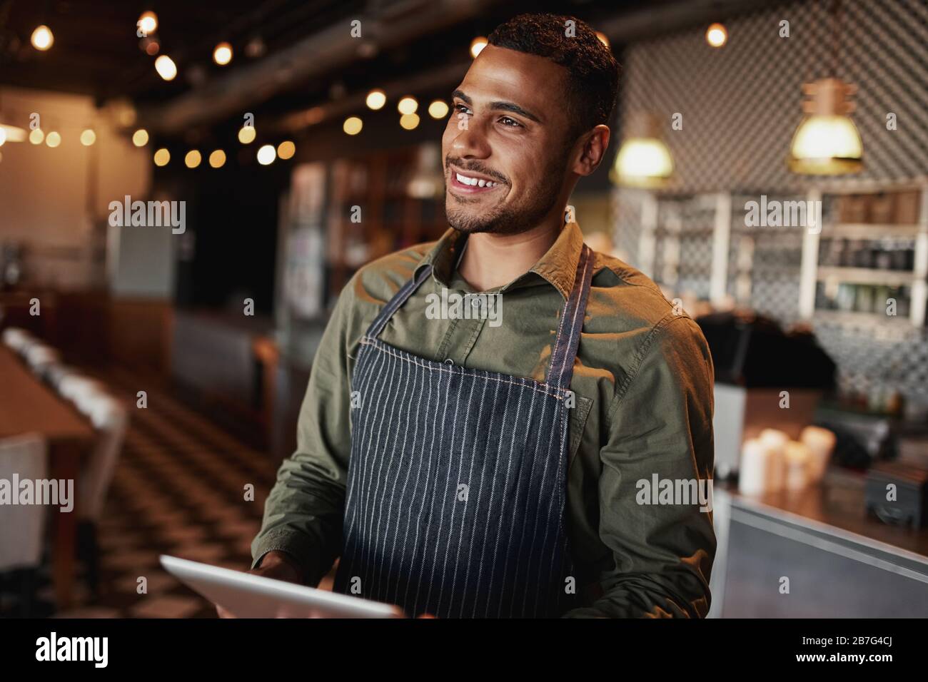 Handsome young man wearing apron holding digital tablet standing in coffee house looking away Stock Photo