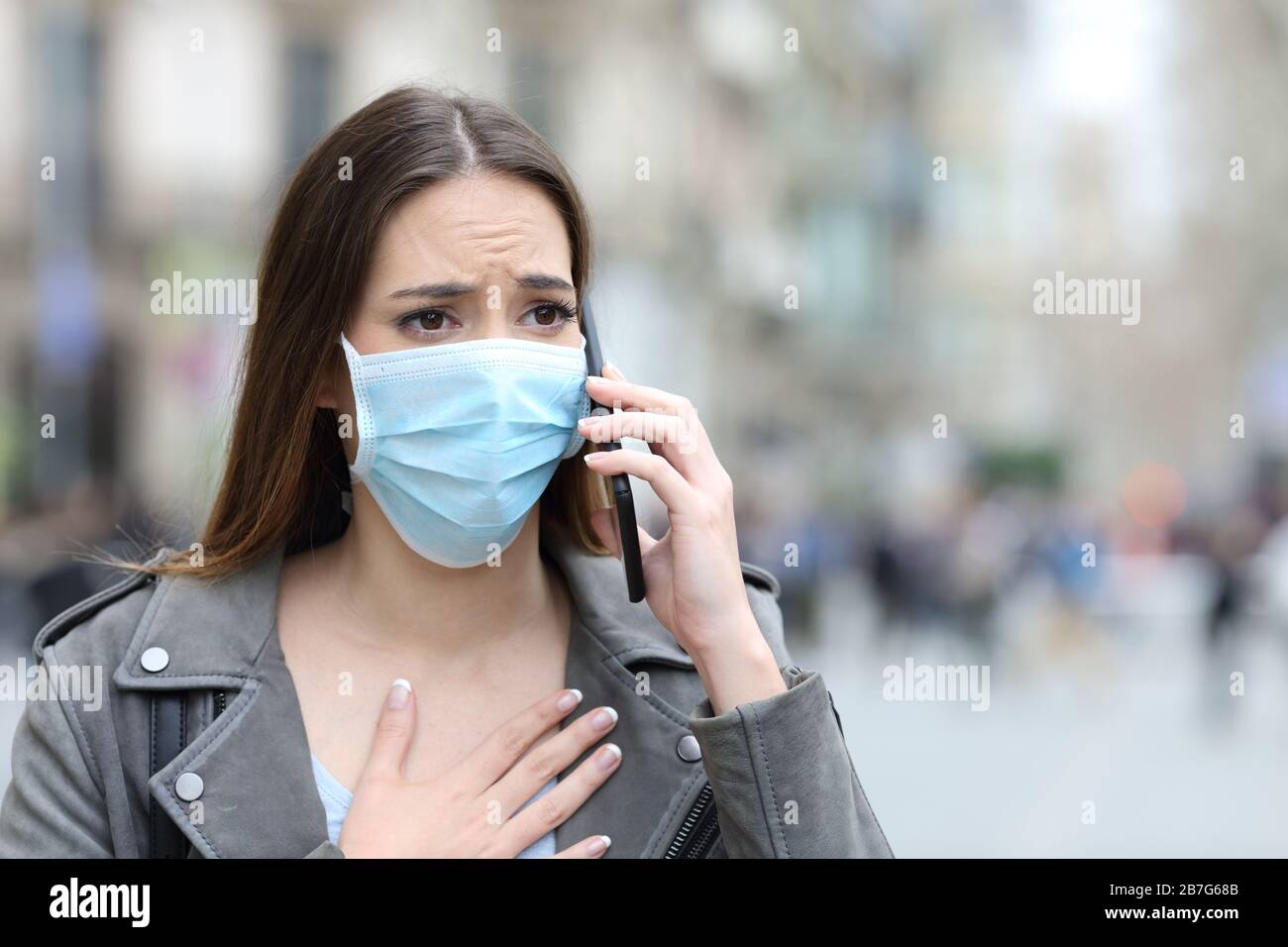 Portrait of a scared girl with hand on chest wearing protective mask calling doctor on smart phone on a city street Stock Photo