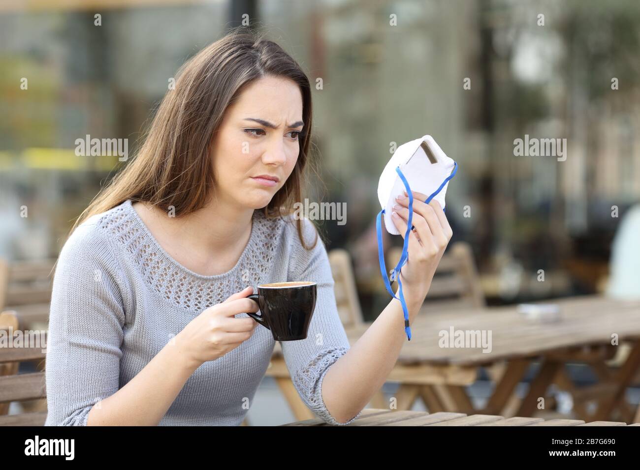 Doubtful young woman looking suspicious at protective face mask on a coffee shop terrace Stock Photo