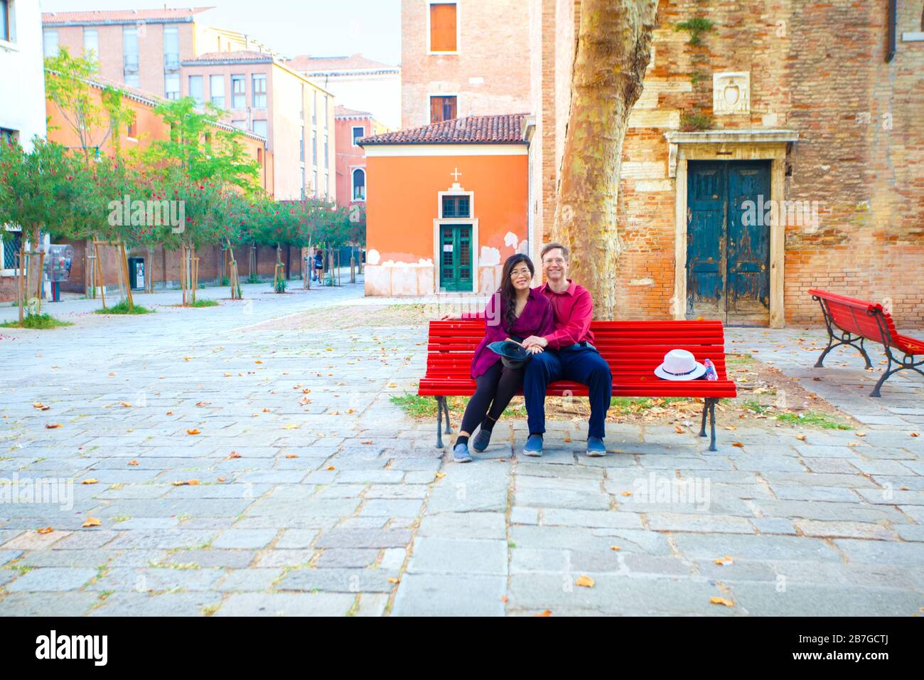 Multiracial tourist couple, Asian Caucasian,  in early fifties sitting together on red bench in empty town square in Venice, Italy Stock Photo