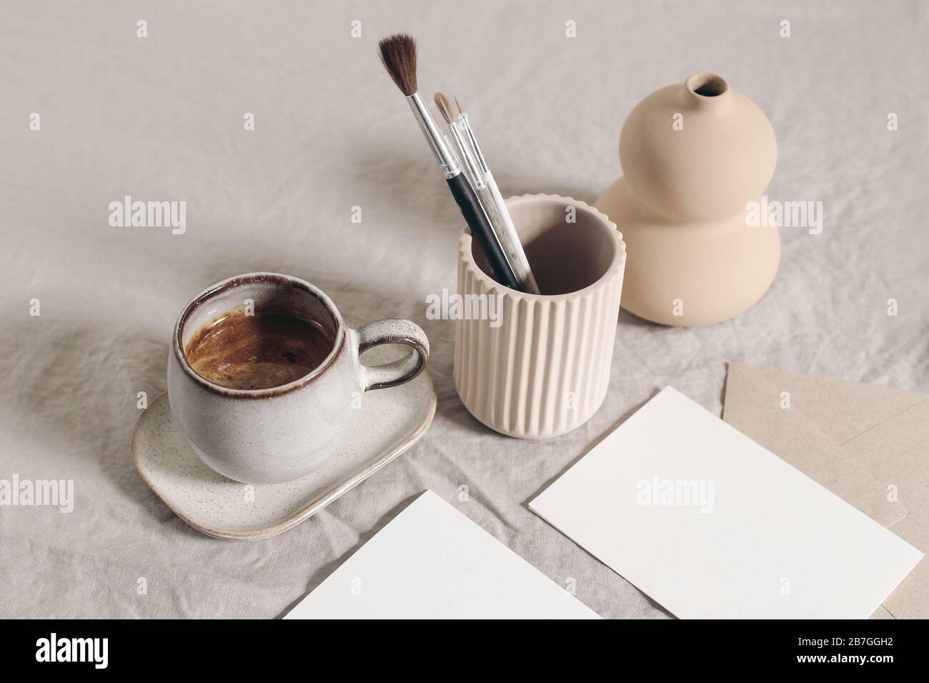 Artistic wokspace, still life. Paint brushes, pencils in ceramic holder, vase, cup of coffee and blank paper card mockups on linen tablecloth. Art Stock Photo